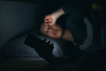 Tired woman rubbing eyes while using mobile phone on bed till late