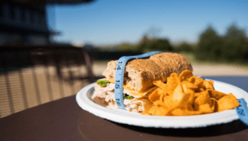 a sandwich and corn chips on a white plate. the sandwich has a tape measure around it