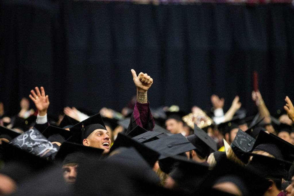 a student standing among graduations in caps and gowns flashes a gig 'em
