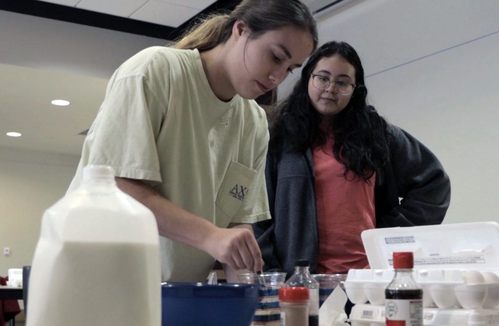 Students crack eggs into a bowl at a table covered with cooking ingredients