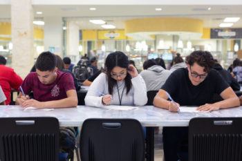 three students look down at papers while sitting at a cafeteria table