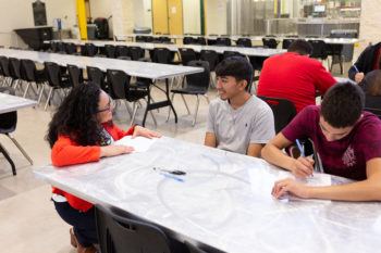 a woman kneels down next two two students sitting at a cafeteria table