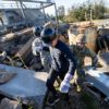 student in black jacket and hard hat walking over earthquake rubble in front of a man in army fatiues