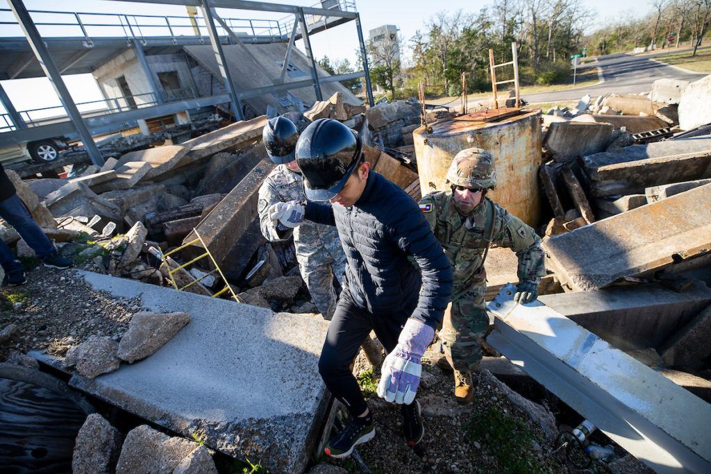 student in black jacket and hard hat walking over earthquake rubble in front of a man in army fatiues