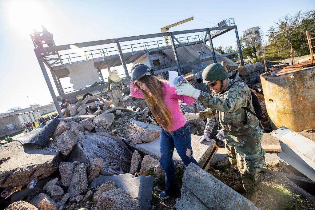 girl wearing pink shirt and hard hat walking over rubble with man in army fatiues