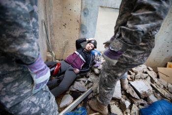 two men in army fatigues standing in front of a student lying in rubble at a mock earthquake site