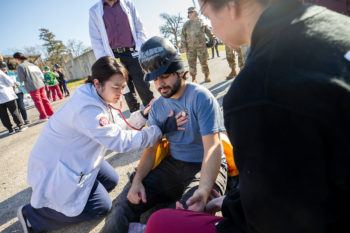 a student in a white doctor's coat listening to the heart of a seated man