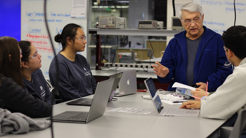 students with laptops sitting at a table listening to a professor talk