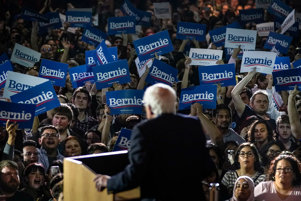 bernie sanders speaking at a lectern in front of a crowd holding signs