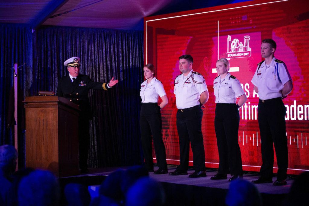 martime academy leader at podium gesturin gto three students in uniform on a stage