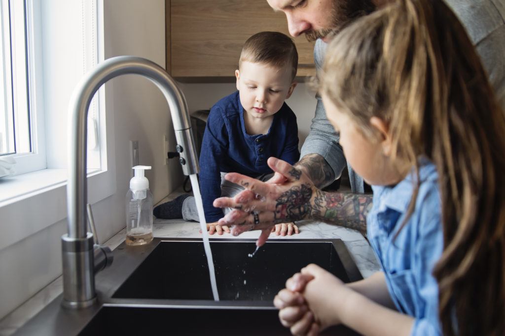 father washing hands at sink while toddler watchers on counter and older child leans on sink