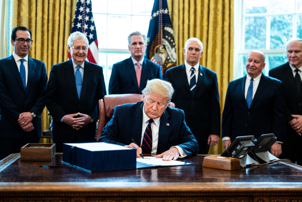 president donald trump seated in the oval office signing bill while cabinet members look on