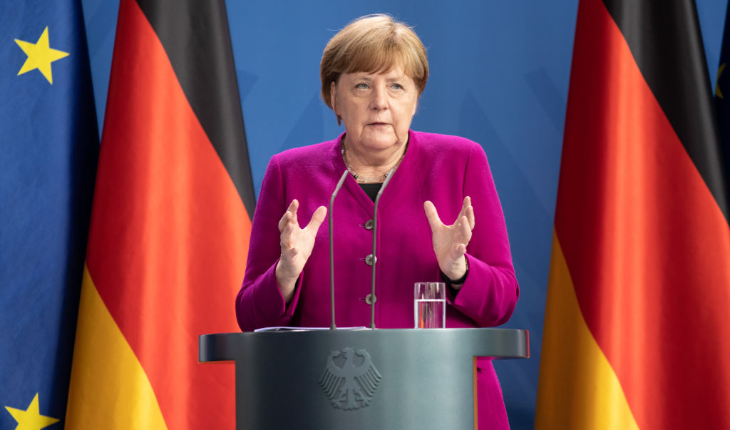 angela merkel speaking behind podium in front of german flags