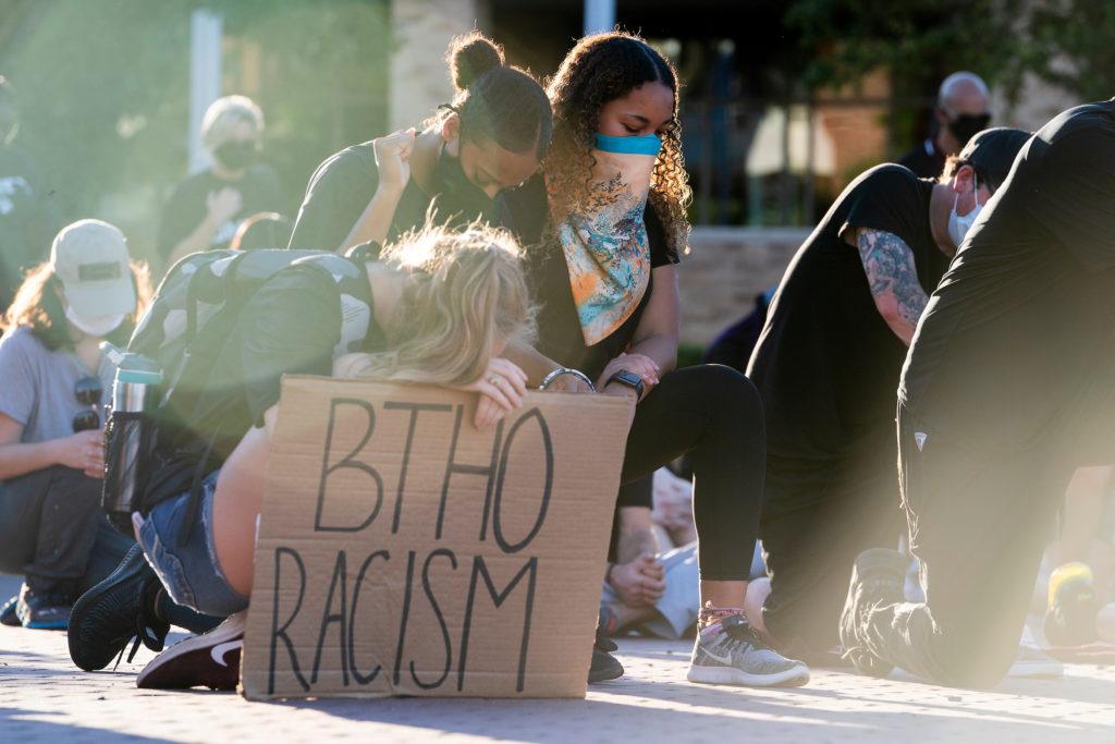 female students kneel together, with one holding a sign that reads BTHO racism