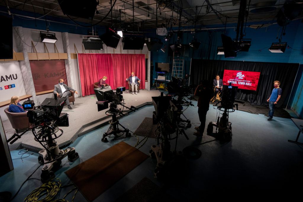 view of cameras and lights pointing toward stage, with three seated peopl being interviewed
