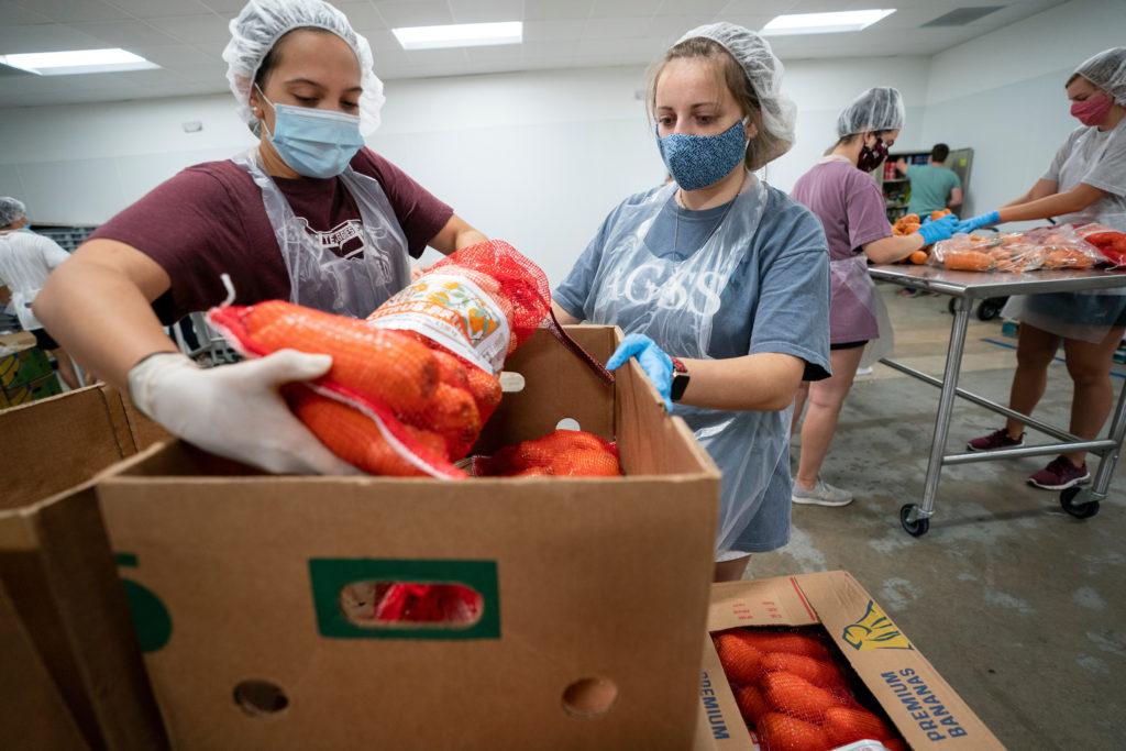two womem wearing face masks and hair nets pick bags of produce up out of cardboard boxes