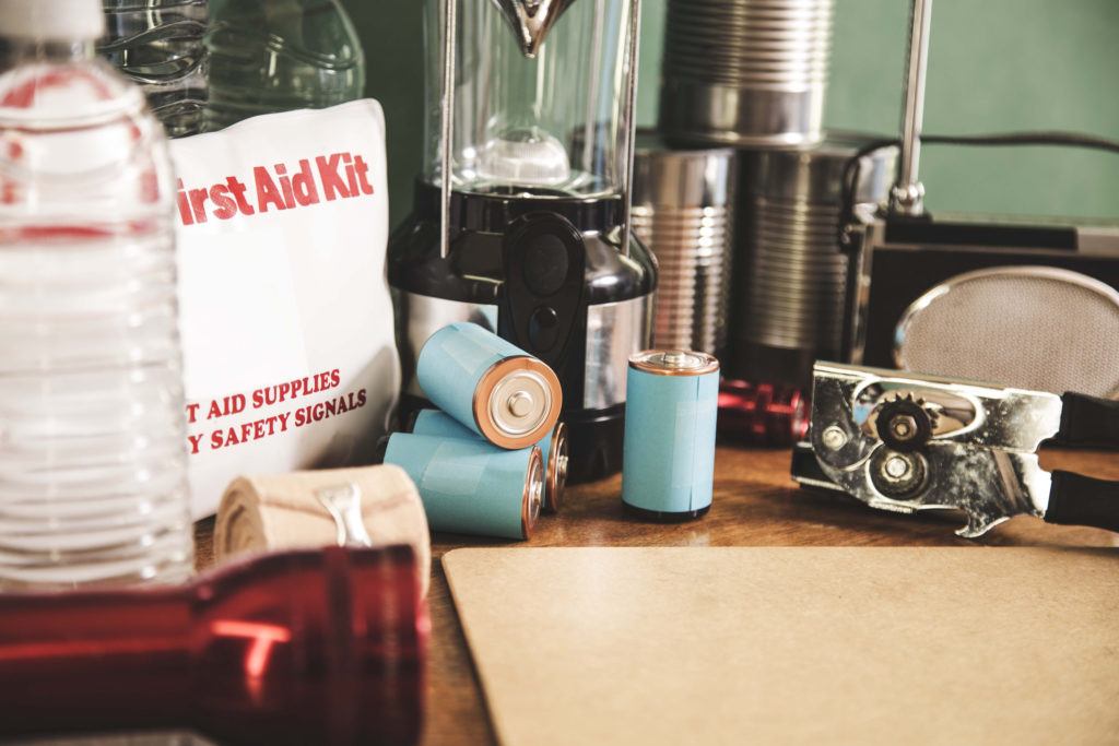 A pile of emergency preparation, natural disaster supplies including: flashlight, first aid kit, lantern, water bottles, canned goods, can opener, radio, backpack, batteries.