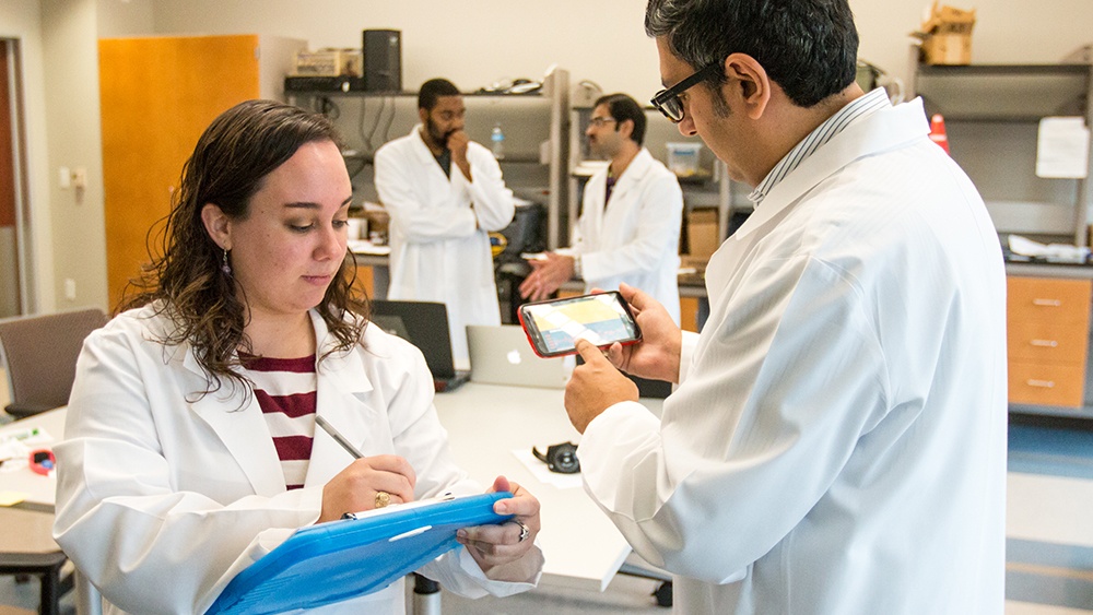 two researchers in white coats work in a lab with two other researchers in the background
