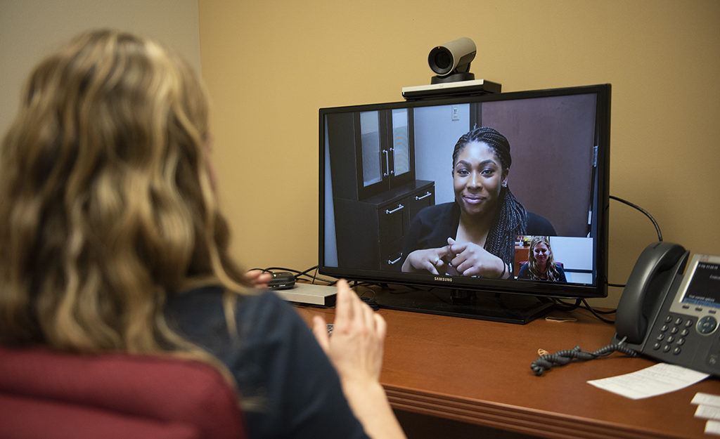 view from behind of a woman sitting at a desk in front of a computer screen displaing another woman speaking into a webcame