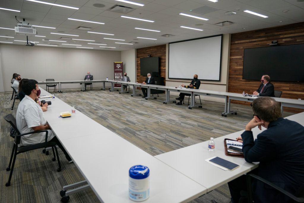 smalll group of people sit several feet apart in a room around a rectangular table