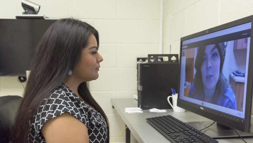 a woman seated at a desk speaks to another woman on a computer screen