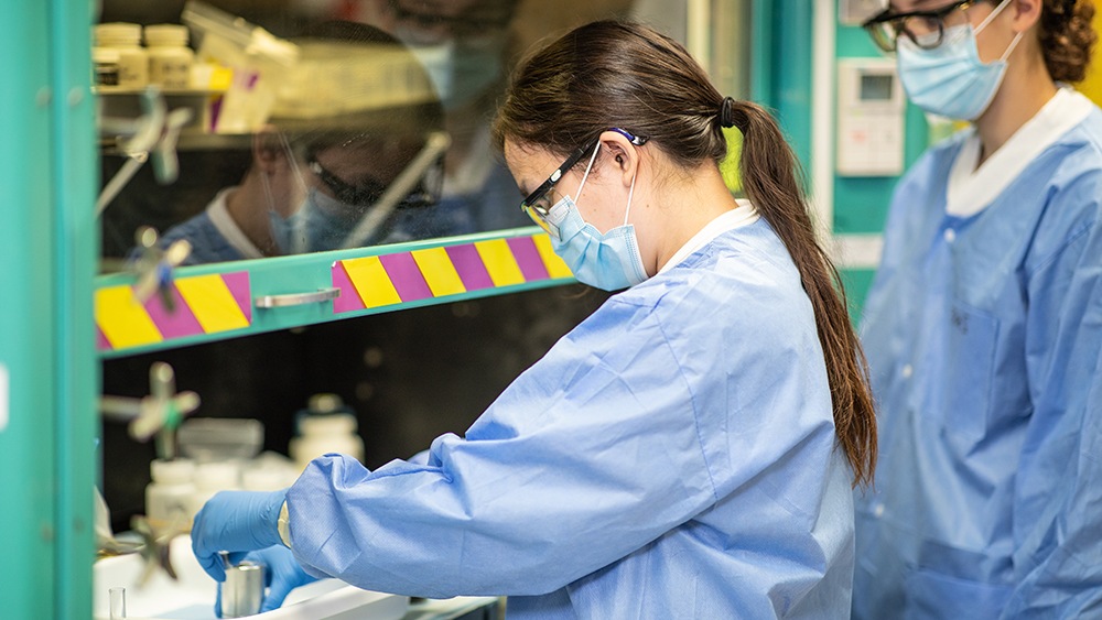 close up of two researchers wearing blue gowns and face coverings working in a lab