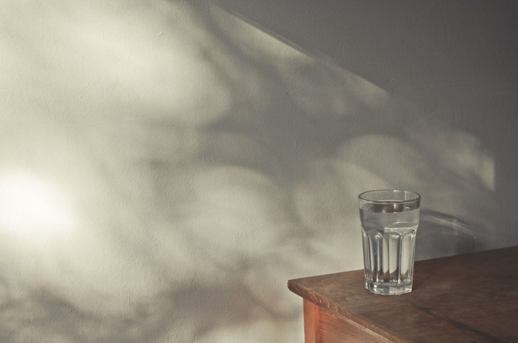 Glass of water on wooden table next to wall with shadows coming from window.