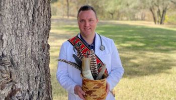 bruce cody standing outside in his white nurse's coat wearing his traditional Native American regalia