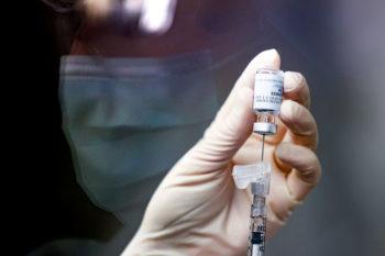 a pharmacy technician is reflected in the glass of a fume hood with her hand in the foreground holding a dose of vaccine