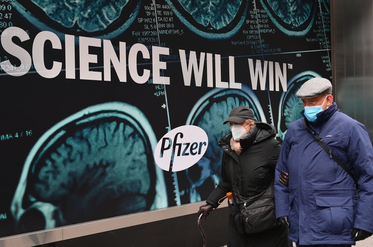 two elderly men walk past building with pfizer advertisement