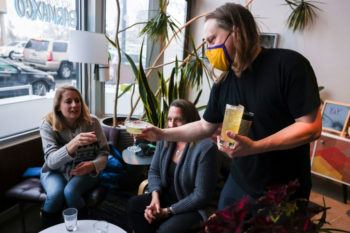 a man wearing a face covering hands a drink to a group of seated women in a bar