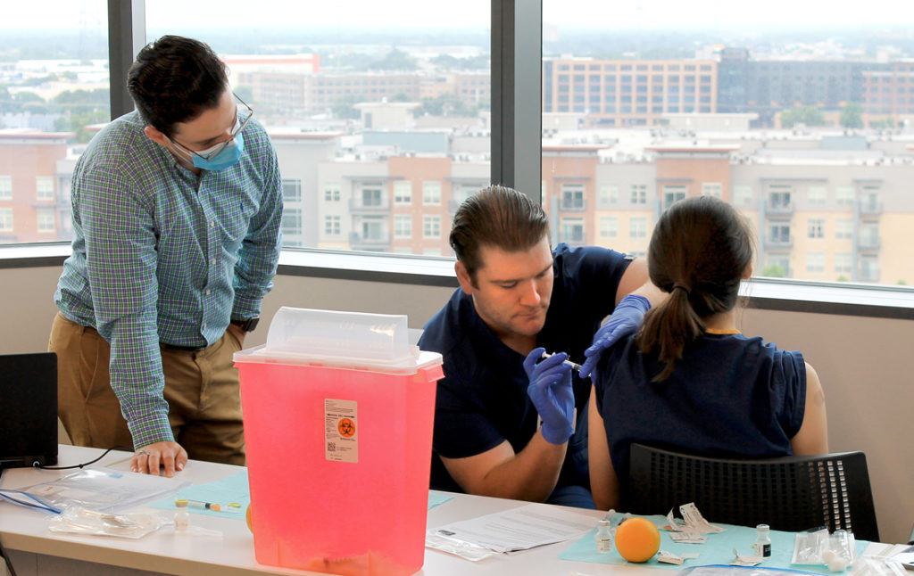 a male student sitting at a table injects a vaccine into another student's arm, as another man looks on