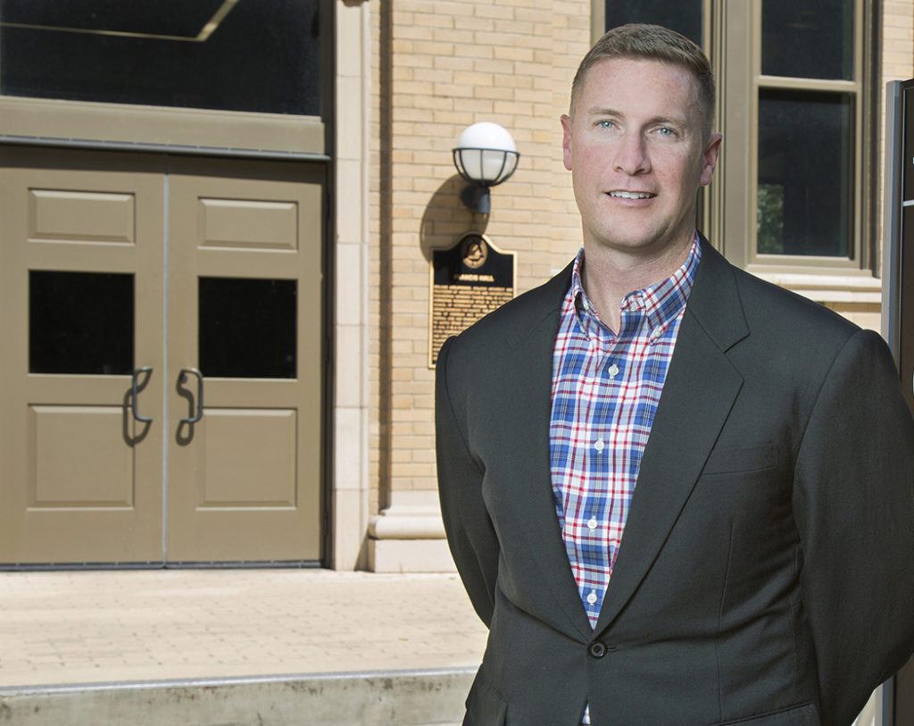 a photo of a man in a dark suit jacket and a red white and blue plaid shirt standing in front of the entrance to Texas A&M's Francis Hall