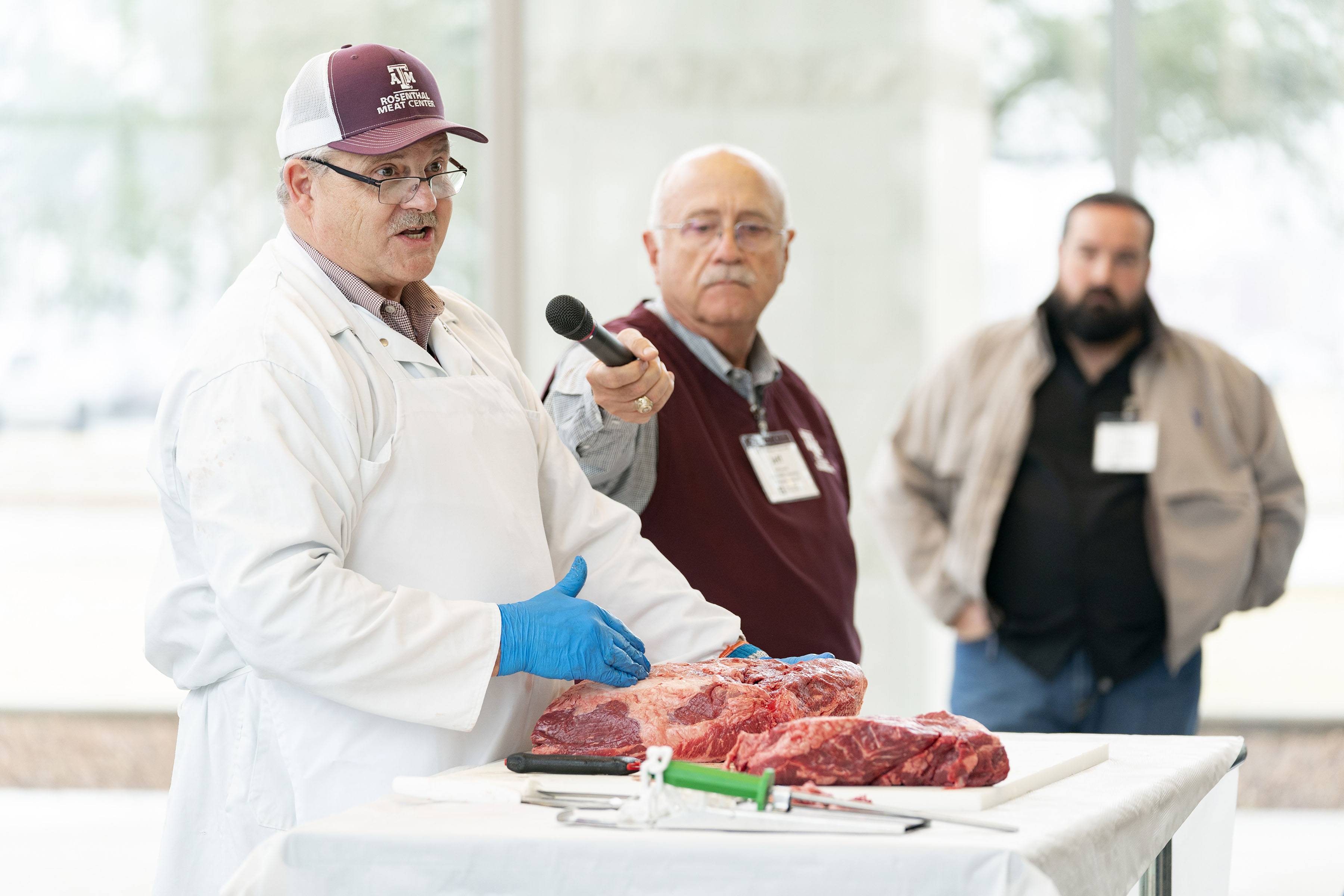 a man wearing a white coat and gloves gives a demonstration using a cut of beef sitting on a table before him