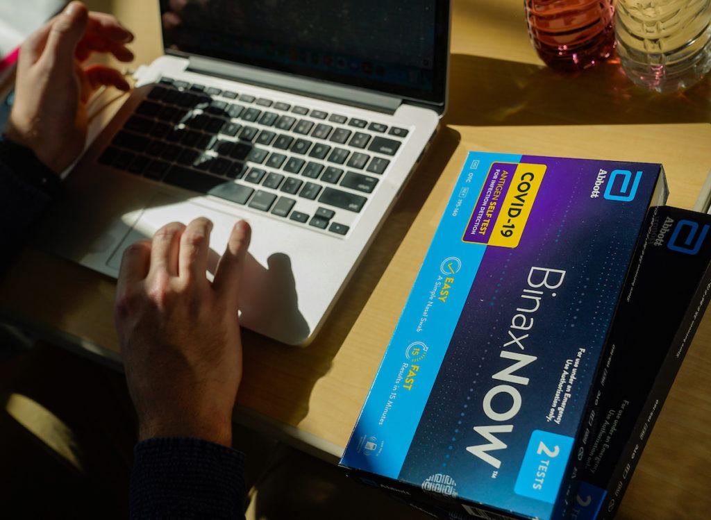 photo of a person typing on a computer with test kit on desk next to them