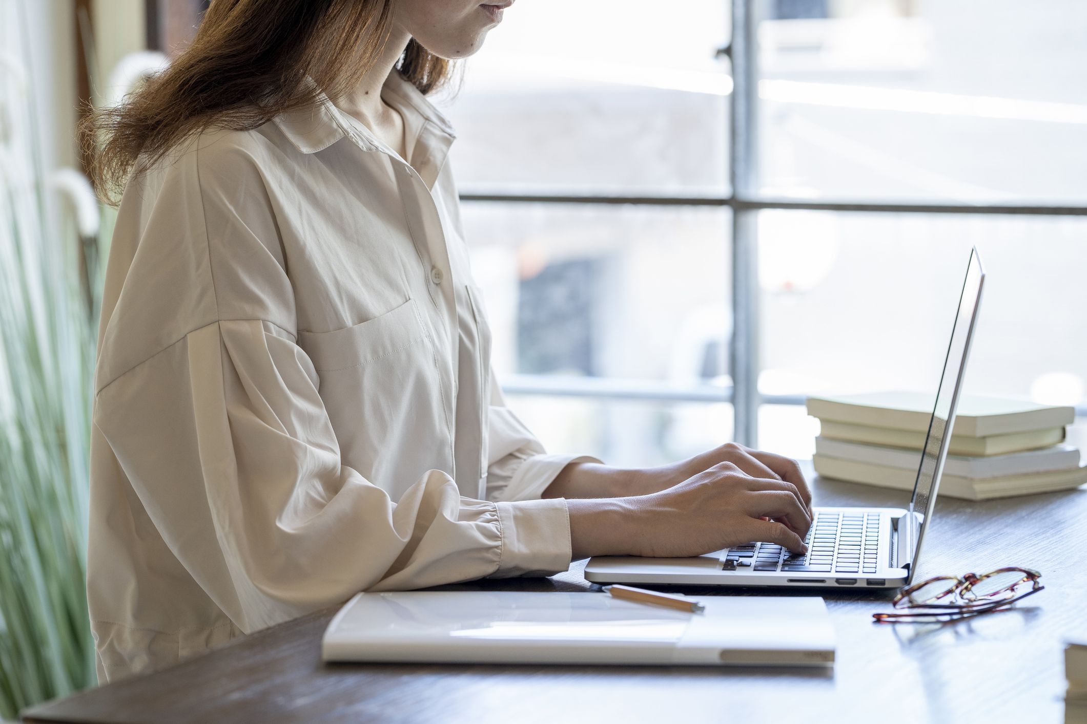 a woman wearing a white shirts sits at a desk at home typing on a computer