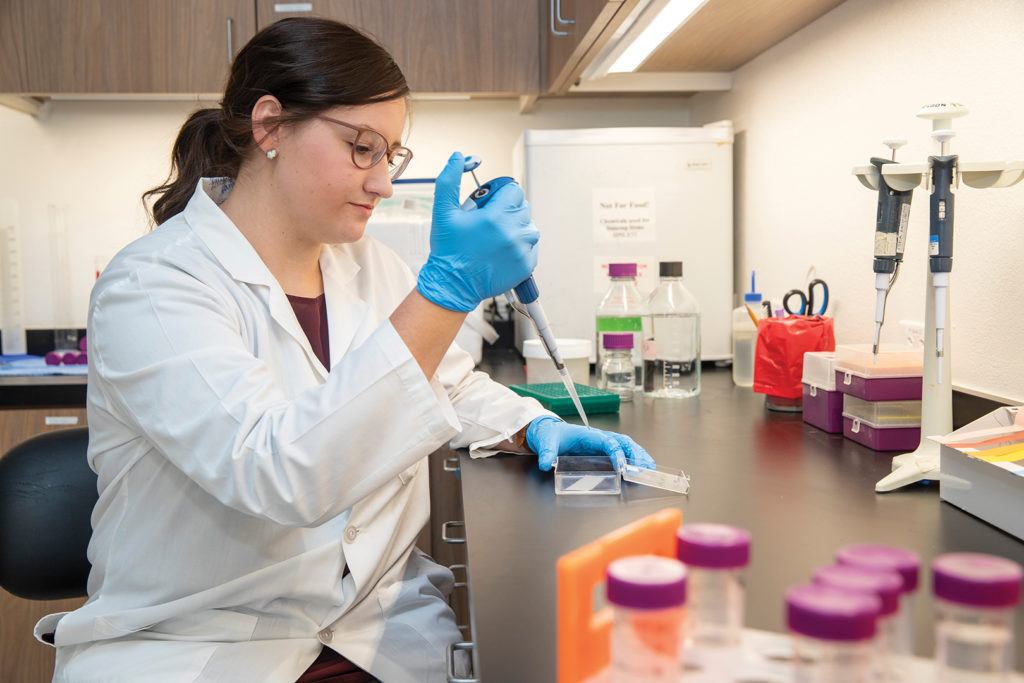 woman wearing a lab coat and goggles sits at a lab bench holding a syringe