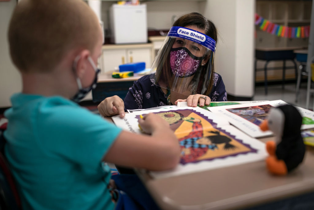 a teaching wearing a face shield kneels next to the desk of a student