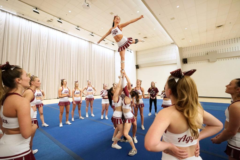 a circle of cheerleaders looks on as a small group of cheerleaders lifts one cheerleader into the air