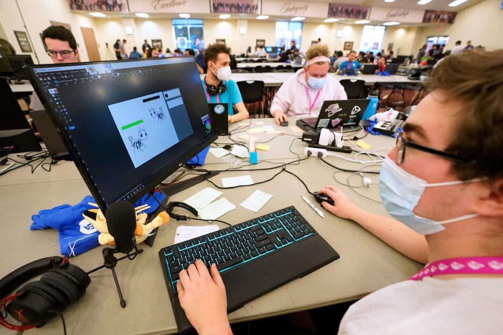a student works at a computer monitor on a video game as he sits at a table surrounded by other students in front of computers