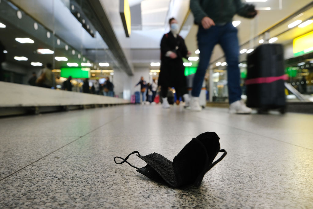 a photo of a black face mask sitting on the floor in the foreground as people with suitcases walk by in the background