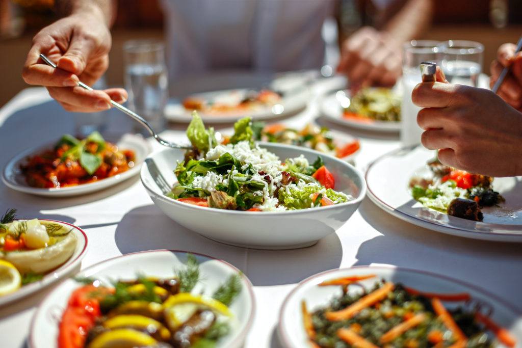 overhead view of a table with plates of food and vegetables, as people eat with forks