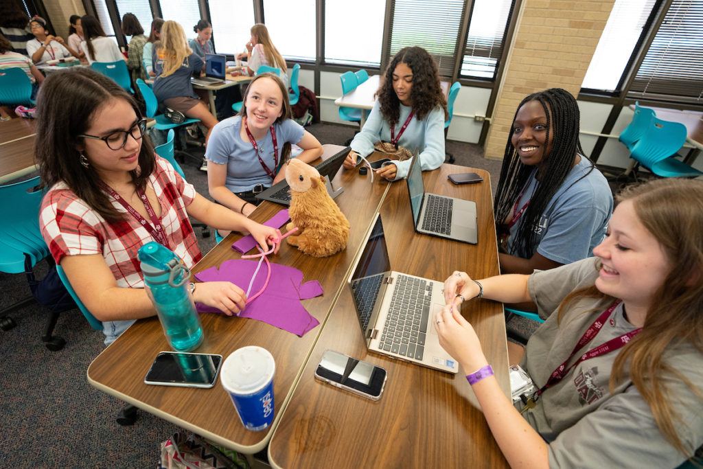 five girls sit around a table working on laptops, with a small stuffed animal sitting in the middle