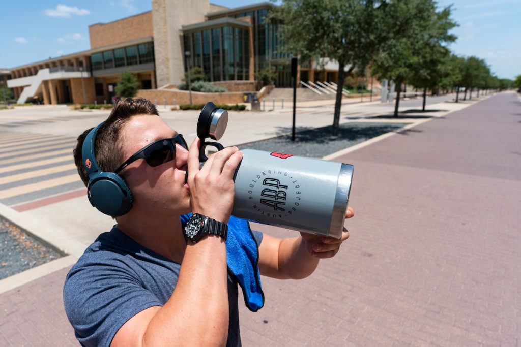 A man on campus wearing sunglasses takes a drink out of a large blue water bottle