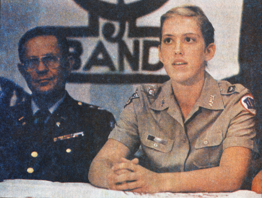 a photo of a young woman in a corps of cadets uniform sitting at a table with an older man wearing glasses and a dark military uniform. behind them is the bottom half of an Aggie Band banner
