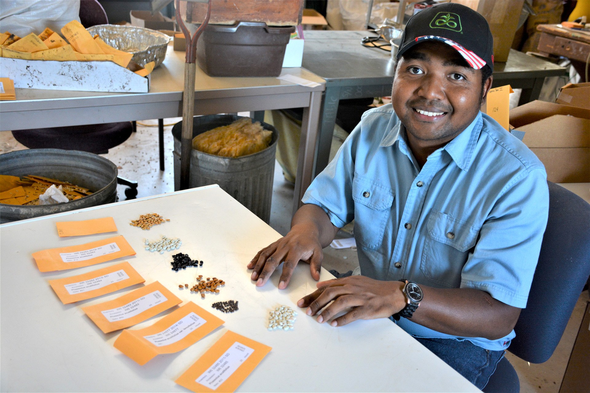 a photo of a man in casual clothes and a cap sitting at a table in front of various piles of different colored beans