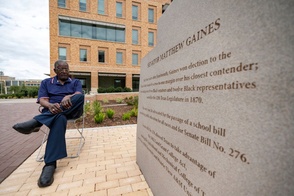 a man seated in a chair looks at the inscription carved on the base of a statue