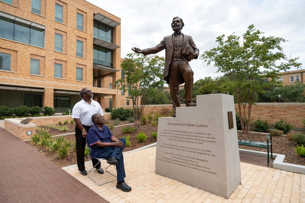 a man seated in a chair, with another man standing behind him, look up at a bronze statue