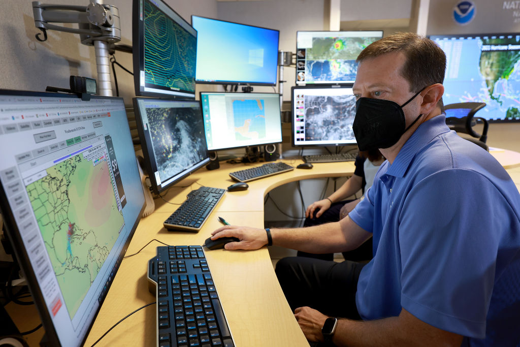a photo of a man in a blue shirt and black facemask sitting at a large desk full of computer monitors. he has his hand on a computer mouse and is looking at a screen showing weather data in the Atlantic ocean.