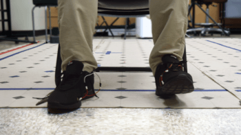 a close-up photo of a person's shins and feet. they are sitting in a chair in a lab wearing a black pair of shoes with various wires sticking out of them.